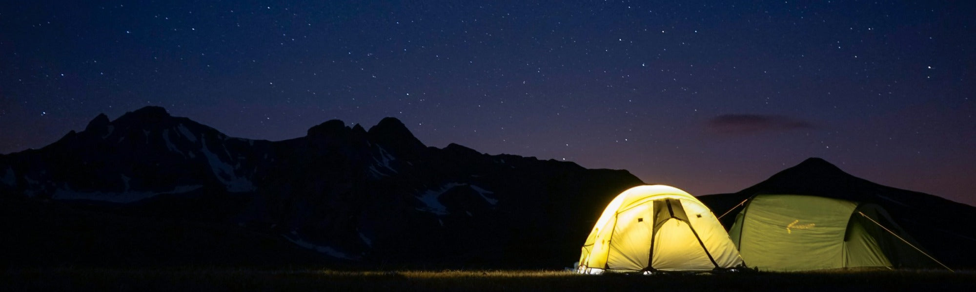 A illuminated yellow tent glowing warmly against a dramatic nighttime mountain landscape with a starry sky